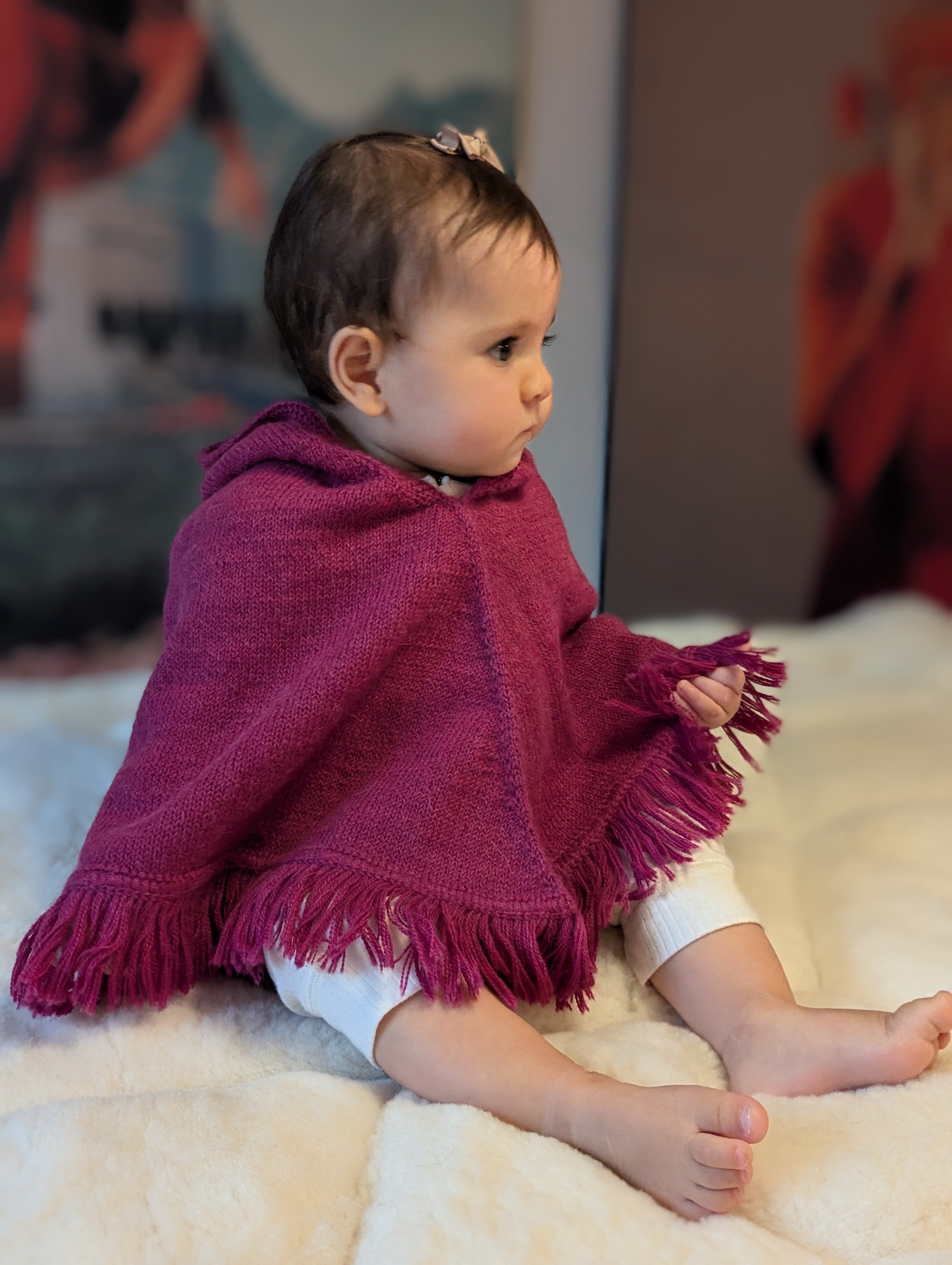 Baby wearing a purple poncho with fringe sitting on a white surface.