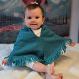 Baby wearing a teal poncho sitting on a white surface with a mountainous background