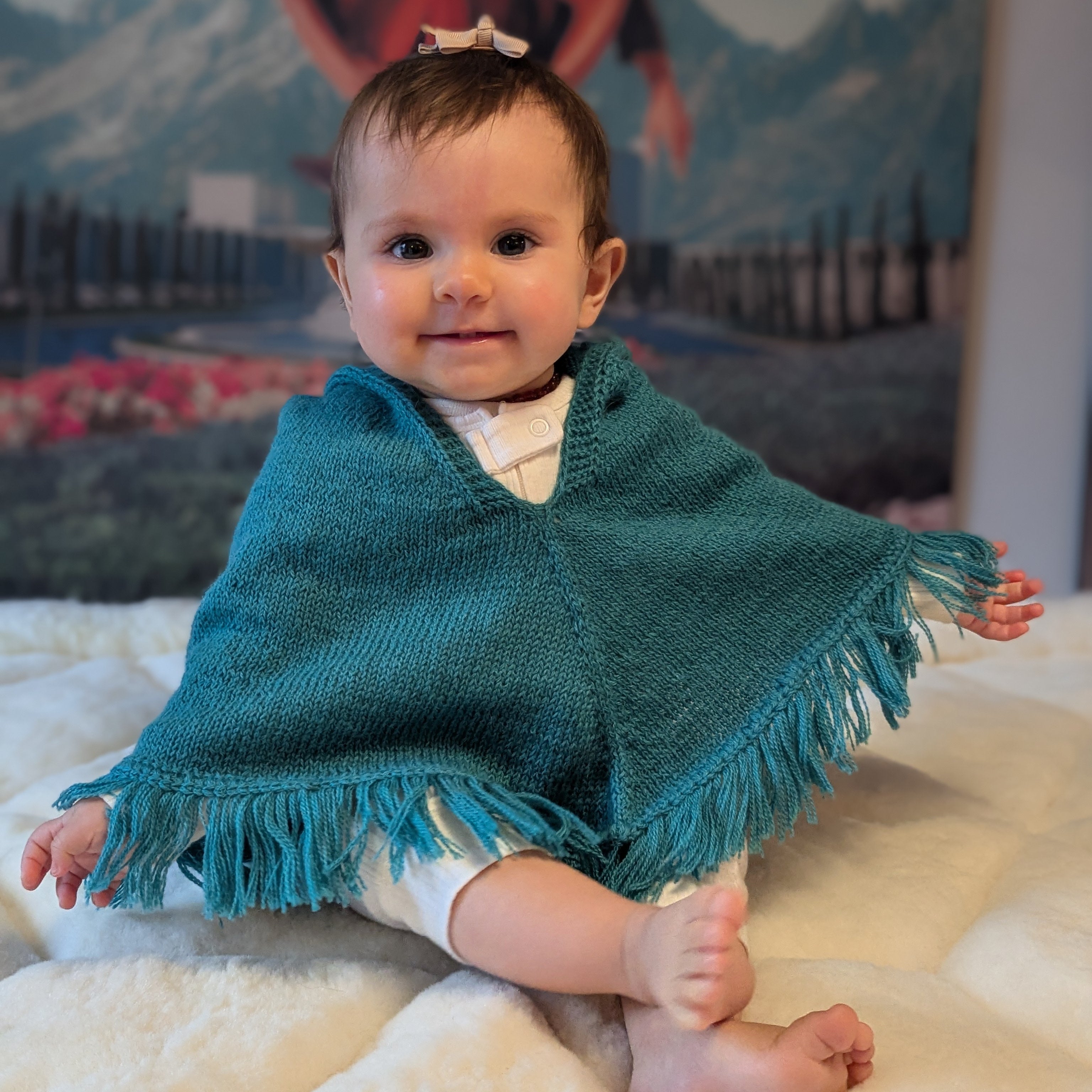 Baby wearing a teal poncho sitting on a white surface with a mountainous background