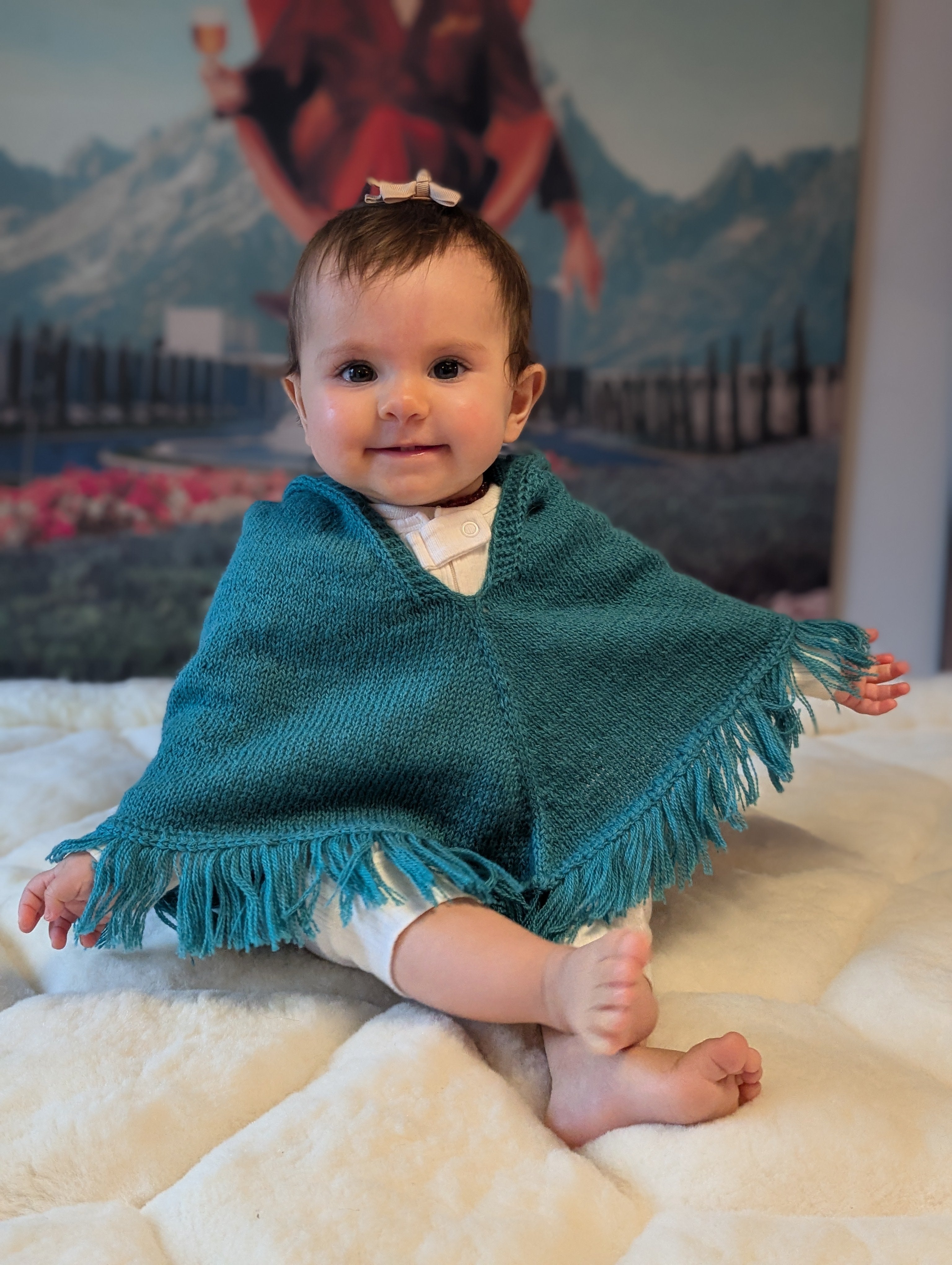 Baby wearing a teal poncho sitting on a white surface with a mountainous background