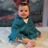 Baby wearing a teal poncho sitting on a white blanket with a mountain scene in the background
