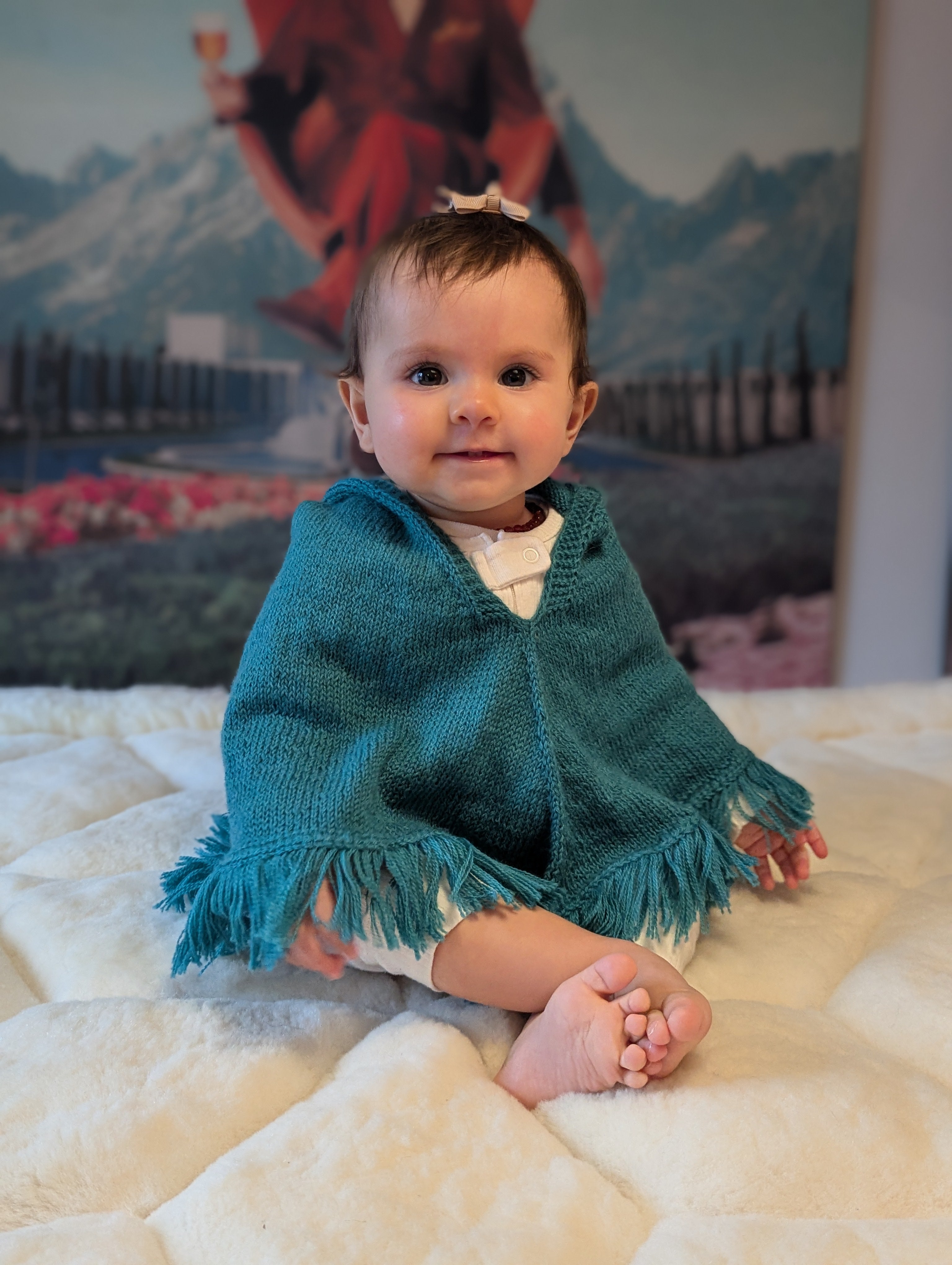 Baby wearing a teal poncho sitting on a white blanket with a mountain scene in the background