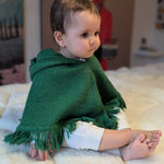Baby wearing a green poncho sitting on a white blanket