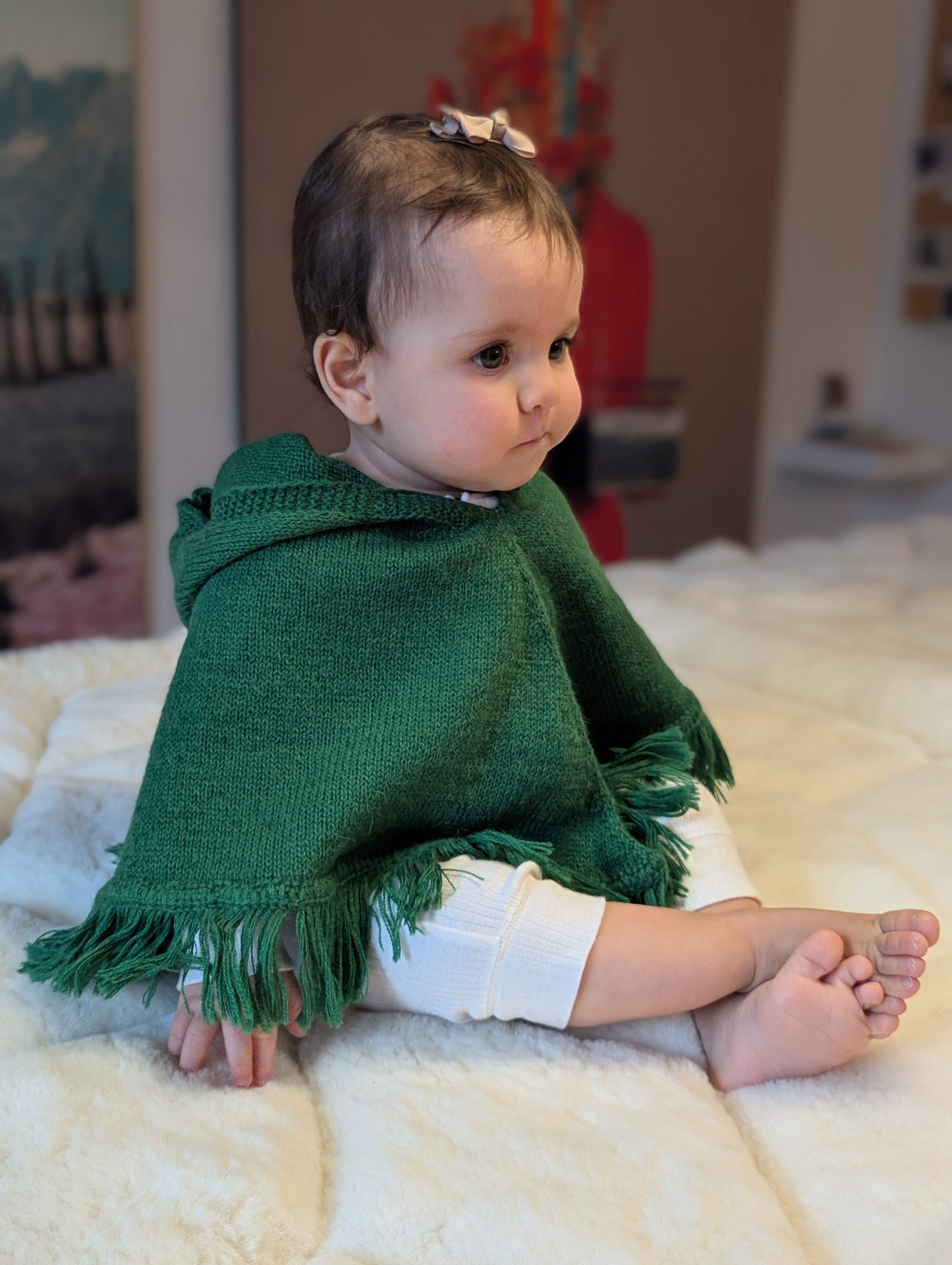 Baby wearing a green poncho sitting on a white blanket