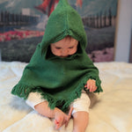 Baby wearing a green hooded cloak on a white blanket with a colorful backdrop