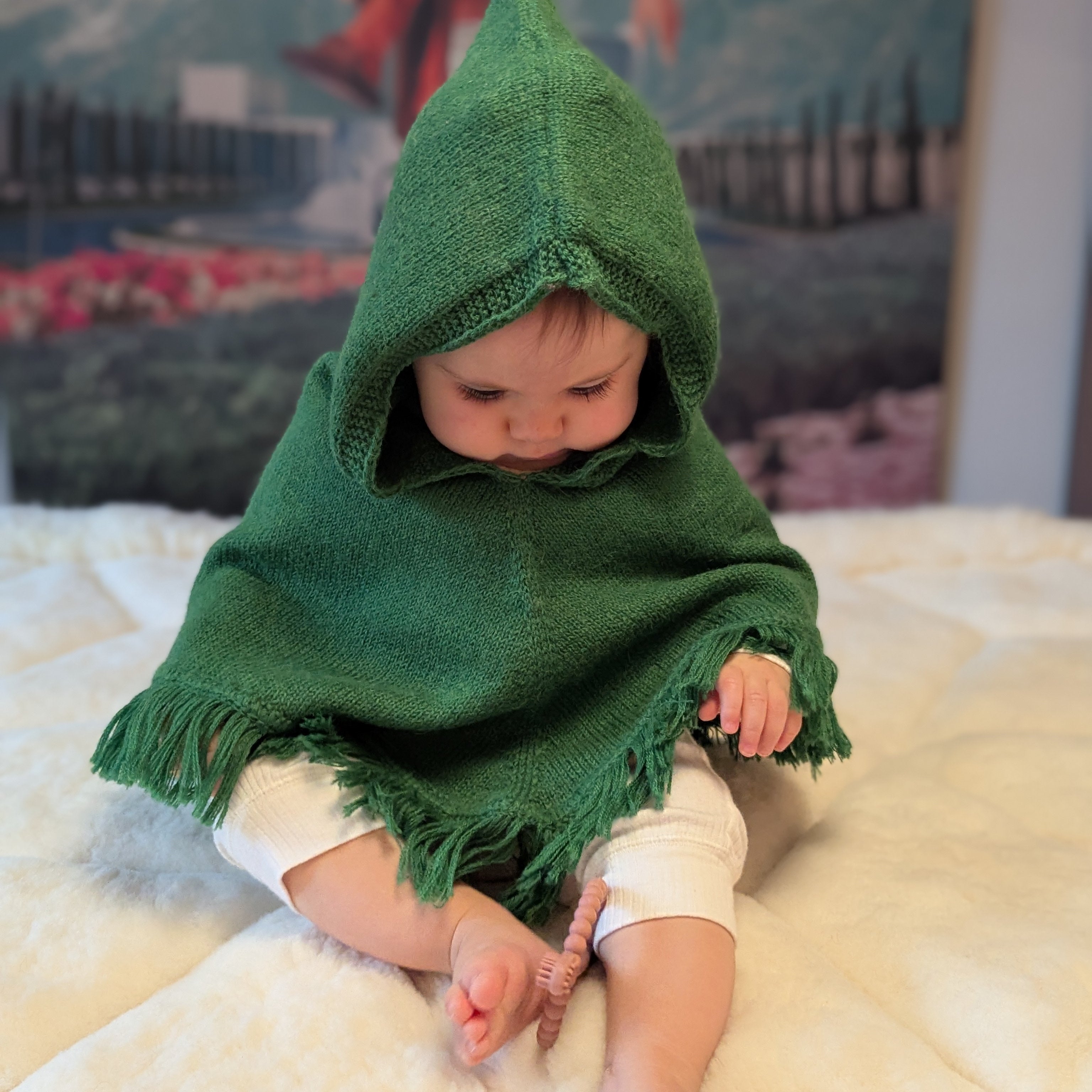 Baby wearing a green hooded cloak on a white blanket with a colorful backdrop