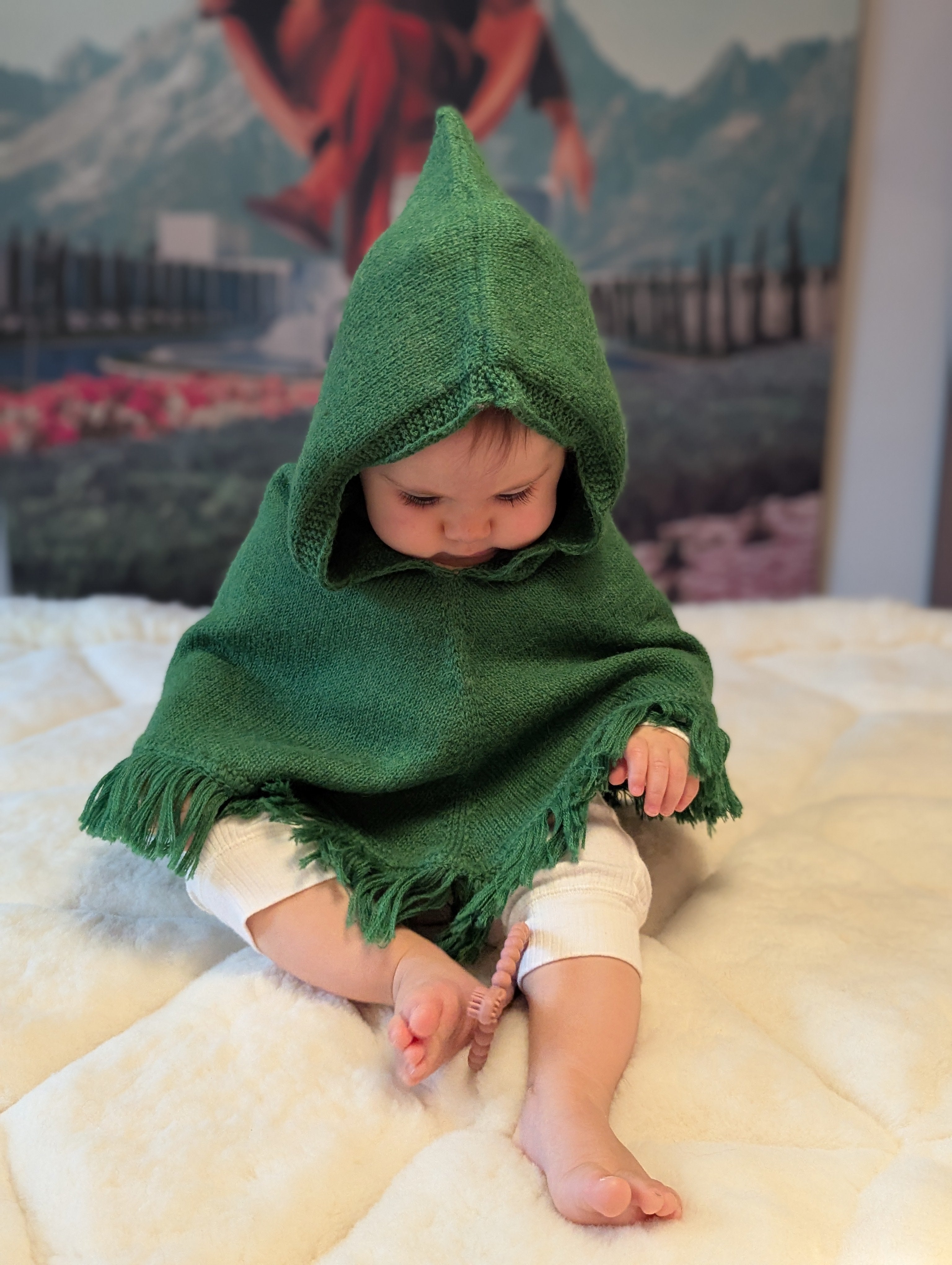 Baby wearing a green hooded cloak on a white blanket with a colorful backdrop