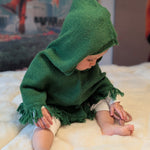 Child wearing a green hooded cloak sitting on a white surface with a colorful poster in the background.