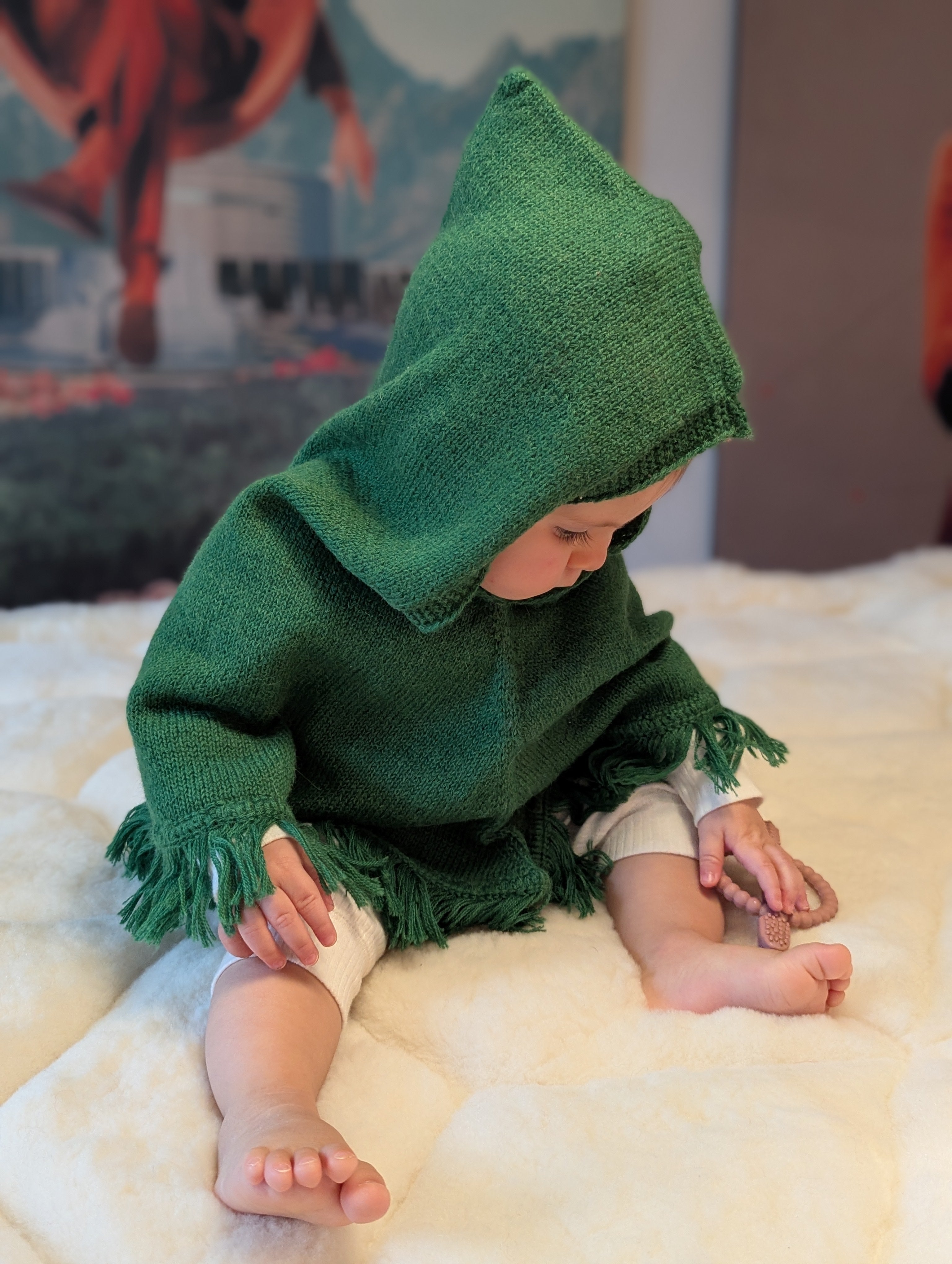 Child wearing a green hooded cloak sitting on a white surface with a colorful poster in the background.