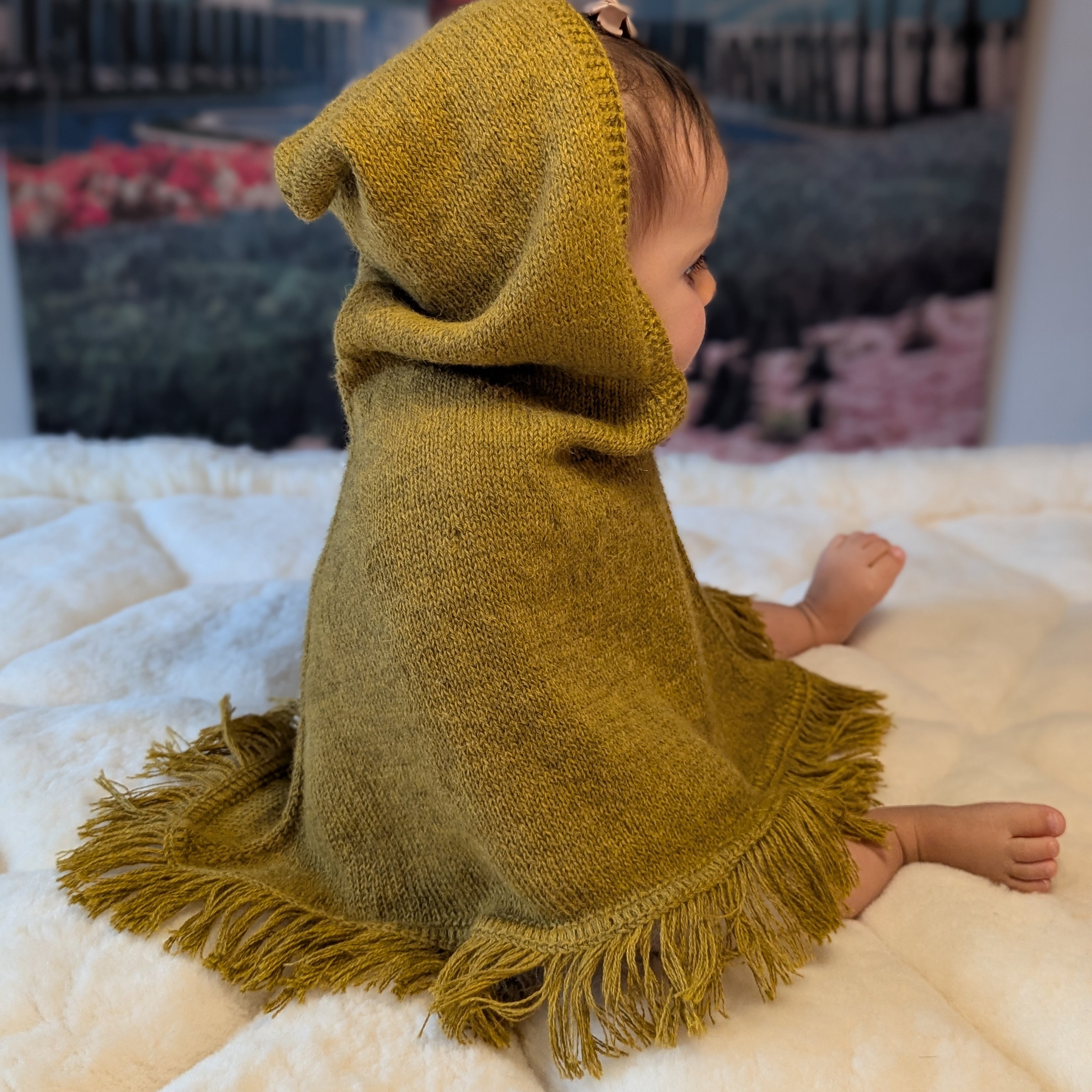 Child wearing a mustard knitted poncho with fringe on a white blanket