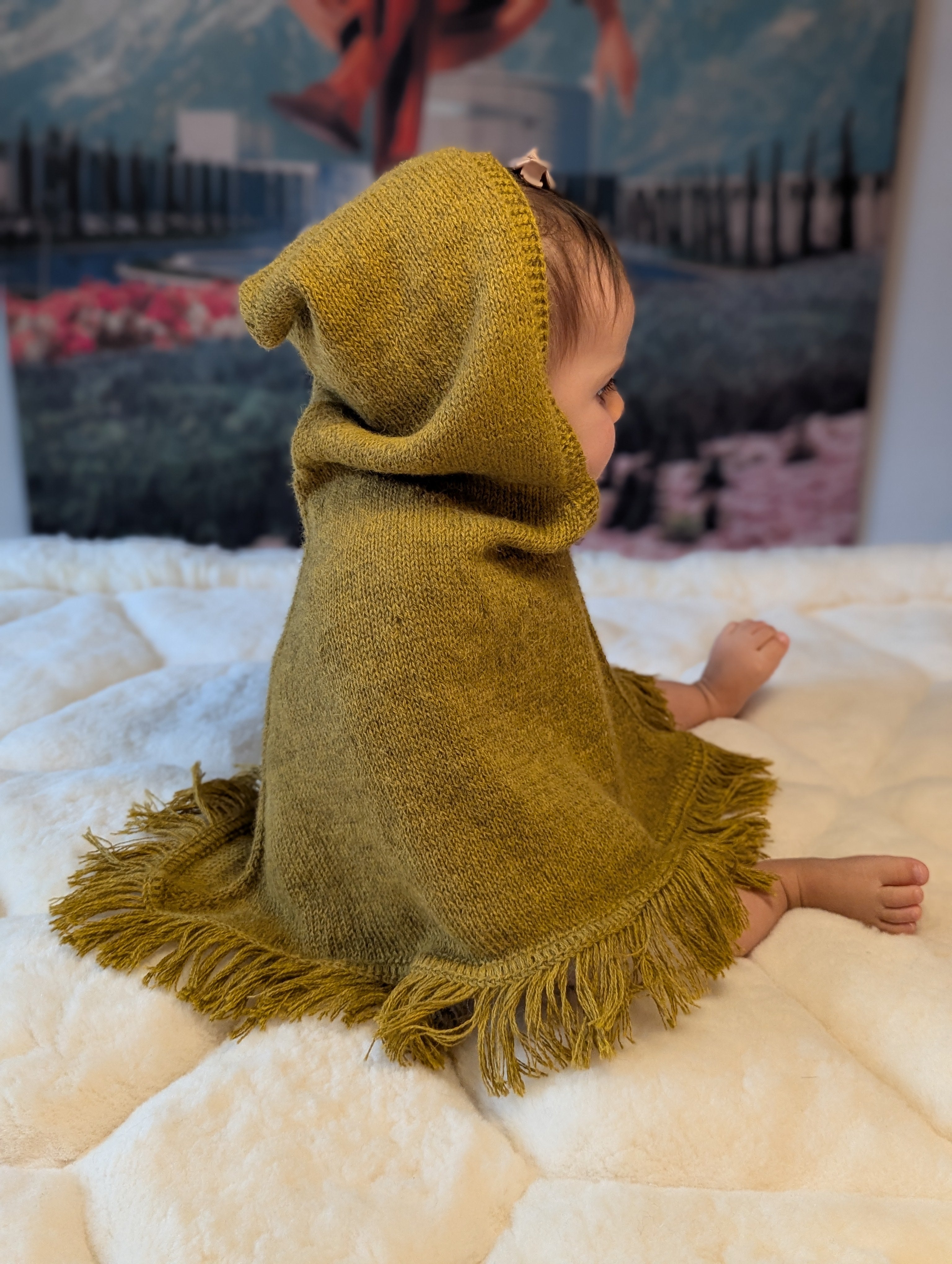 Child wearing a mustard knitted poncho with fringe on a white blanket