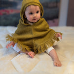 Baby wearing a mustard hooded poncho sitting on a white surface with a blurred background