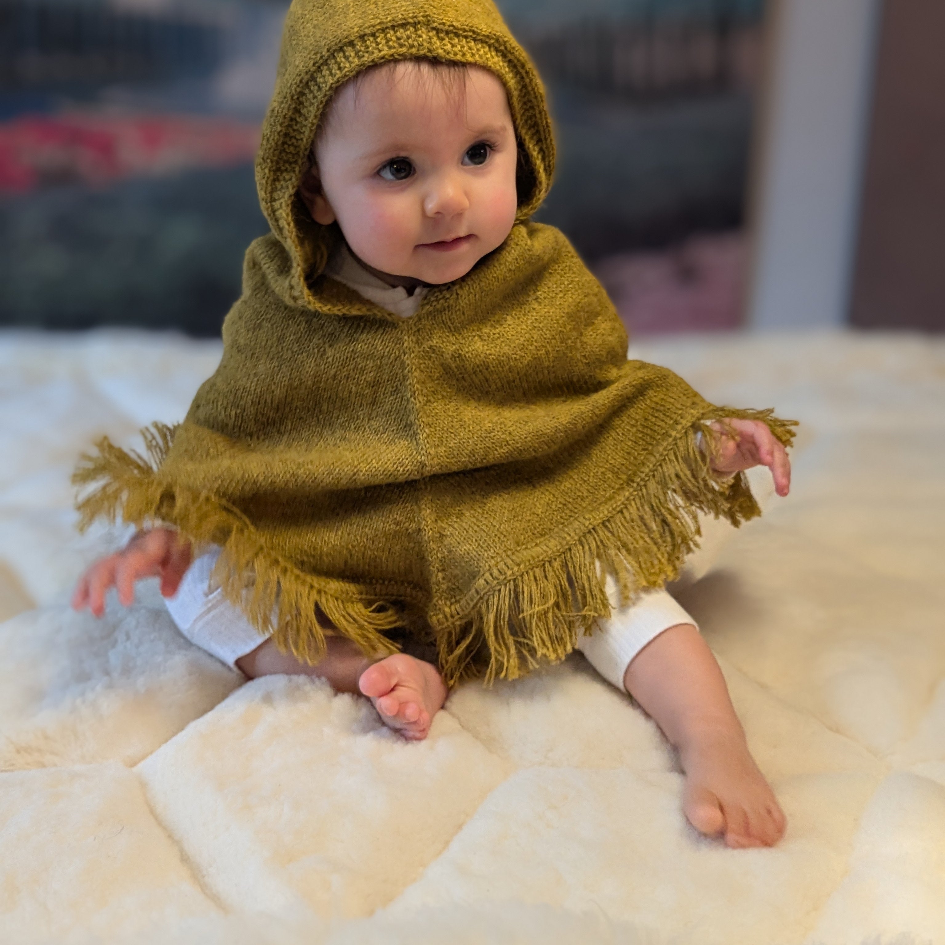 Baby wearing a mustard hooded poncho sitting on a white surface with a blurred background