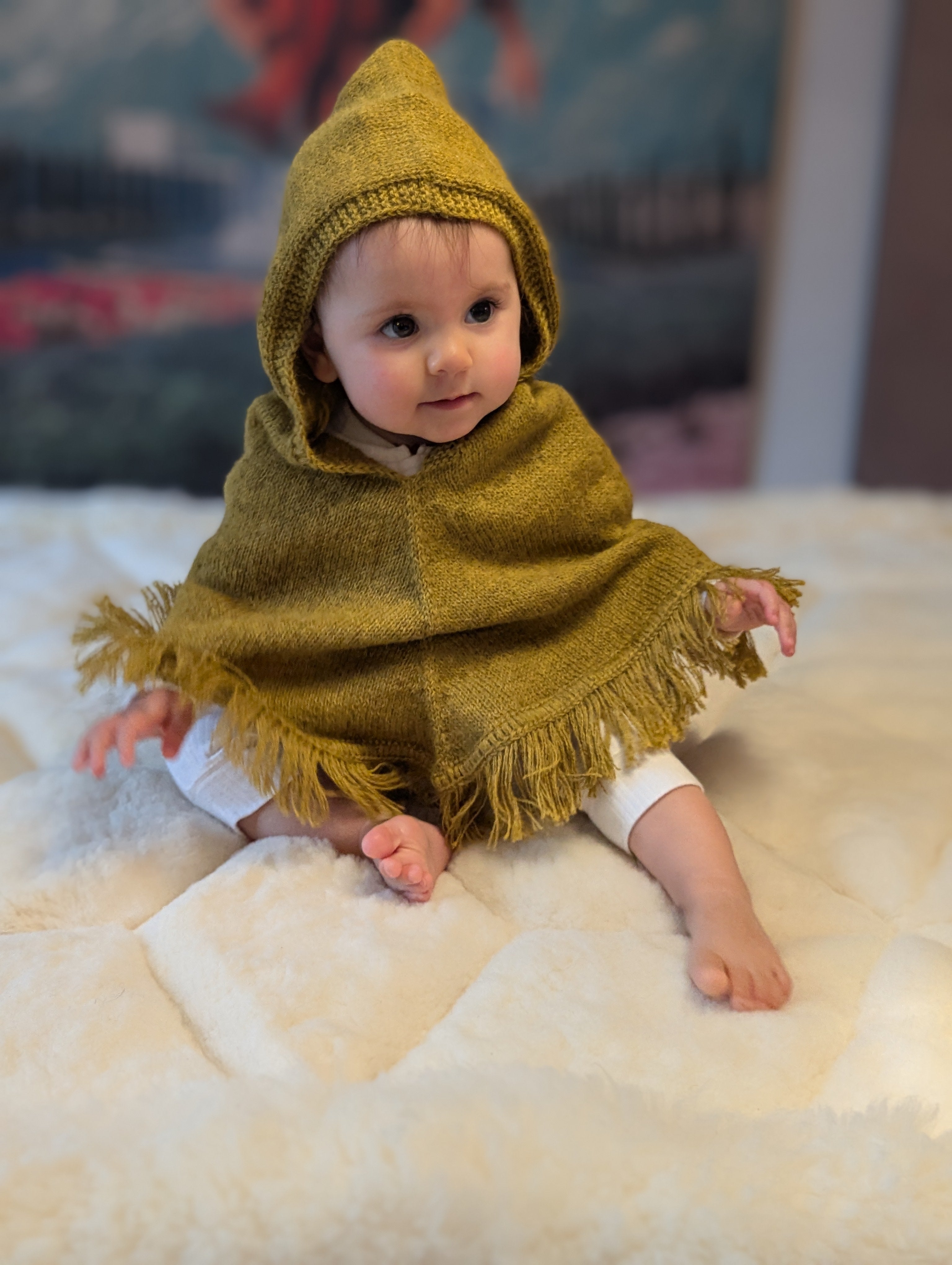 Baby wearing a mustard hooded poncho sitting on a white surface with a blurred background
