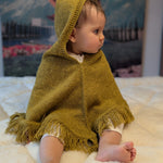 Child wearing a mustard hooded poncho sitting on a white surface with a mountainous landscape in the background.