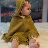 Child wearing a mustard hooded poncho sitting on a white surface with a mountainous landscape in the background.
