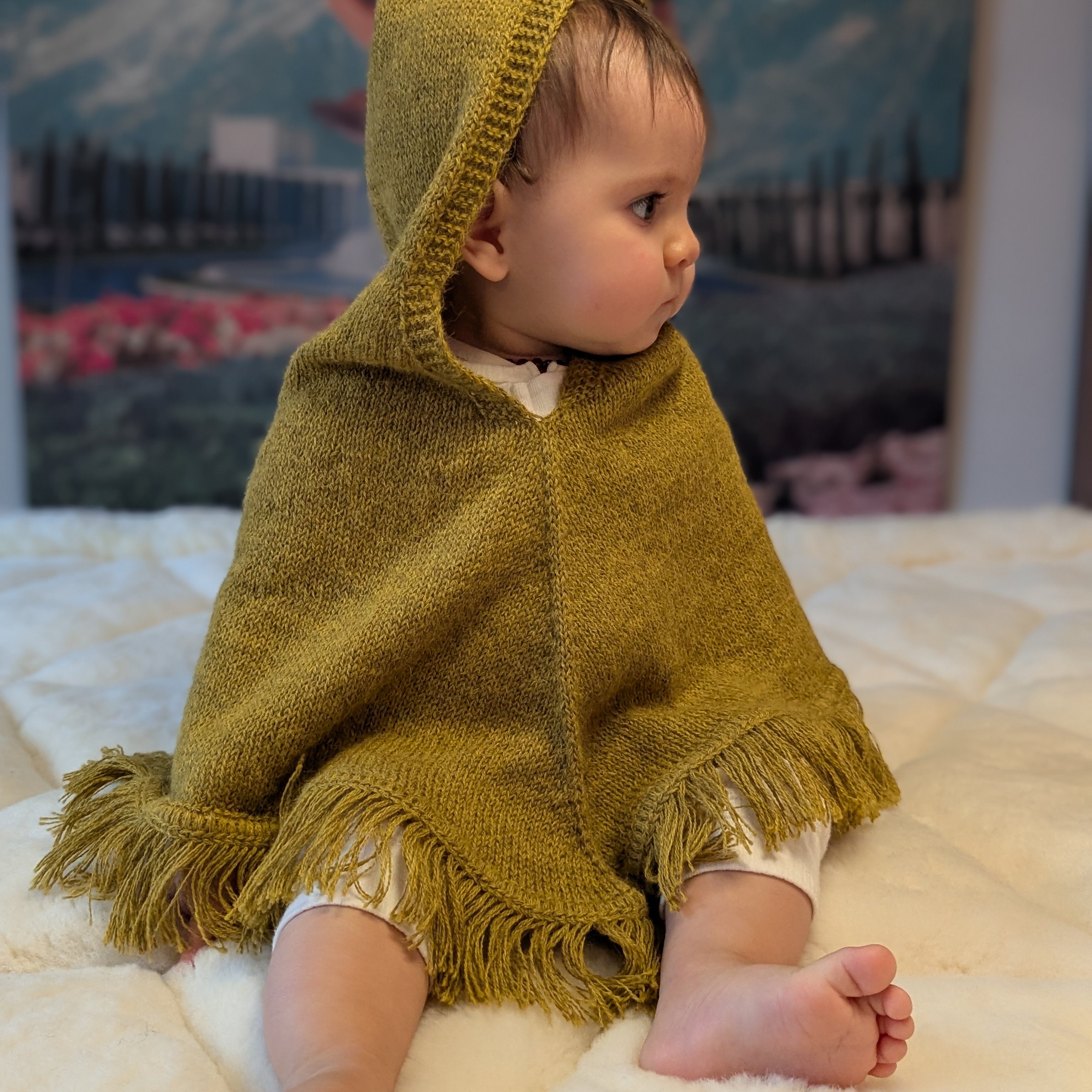 Child wearing a mustard hooded poncho sitting on a white surface with a mountainous landscape in the background.