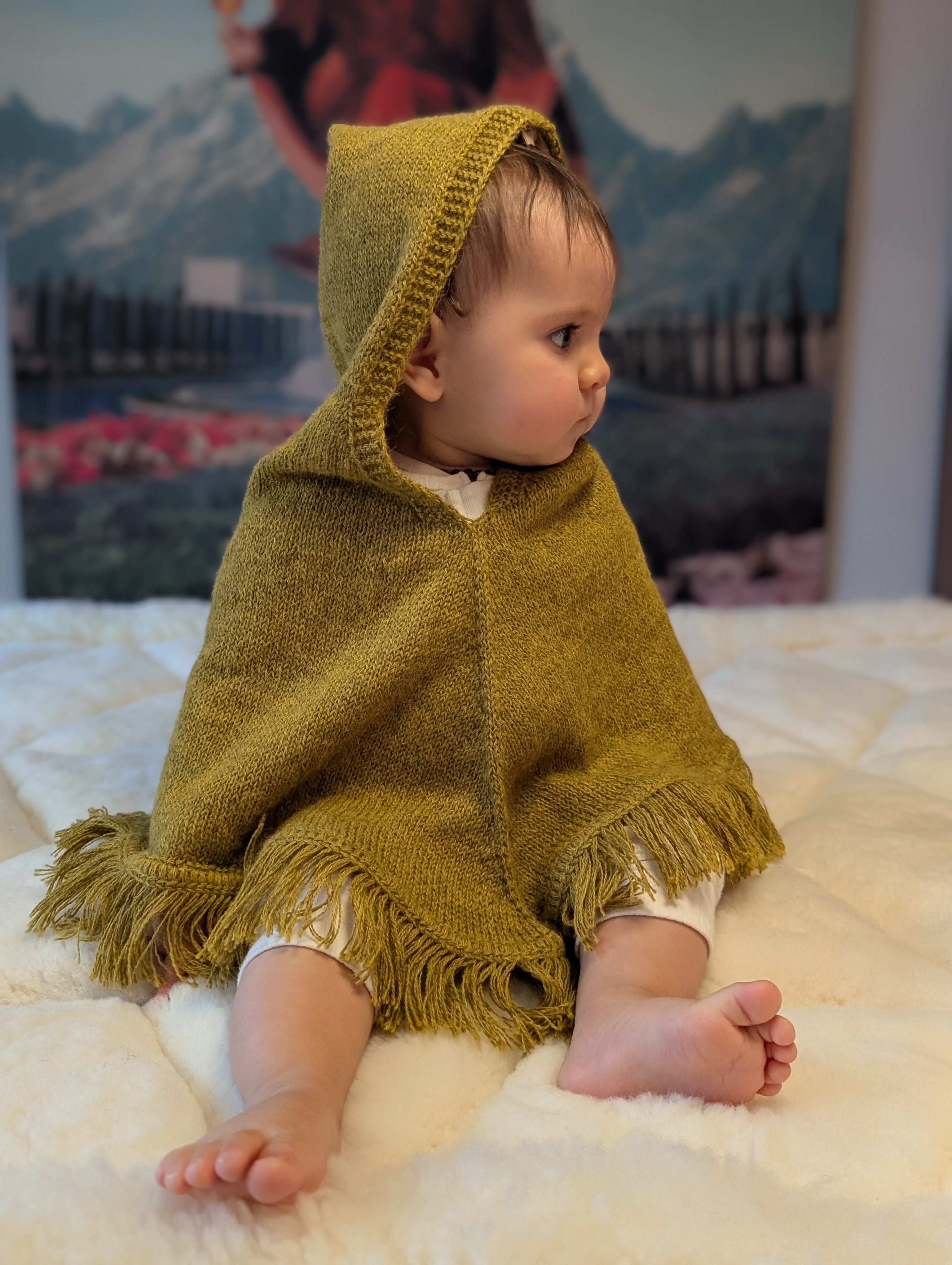 Child wearing a mustard hooded poncho sitting on a white surface with a mountainous landscape in the background.