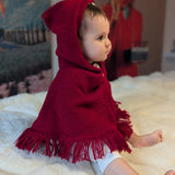 Baby wearing a red hooded cloak sitting on a white surface with a colorful background.