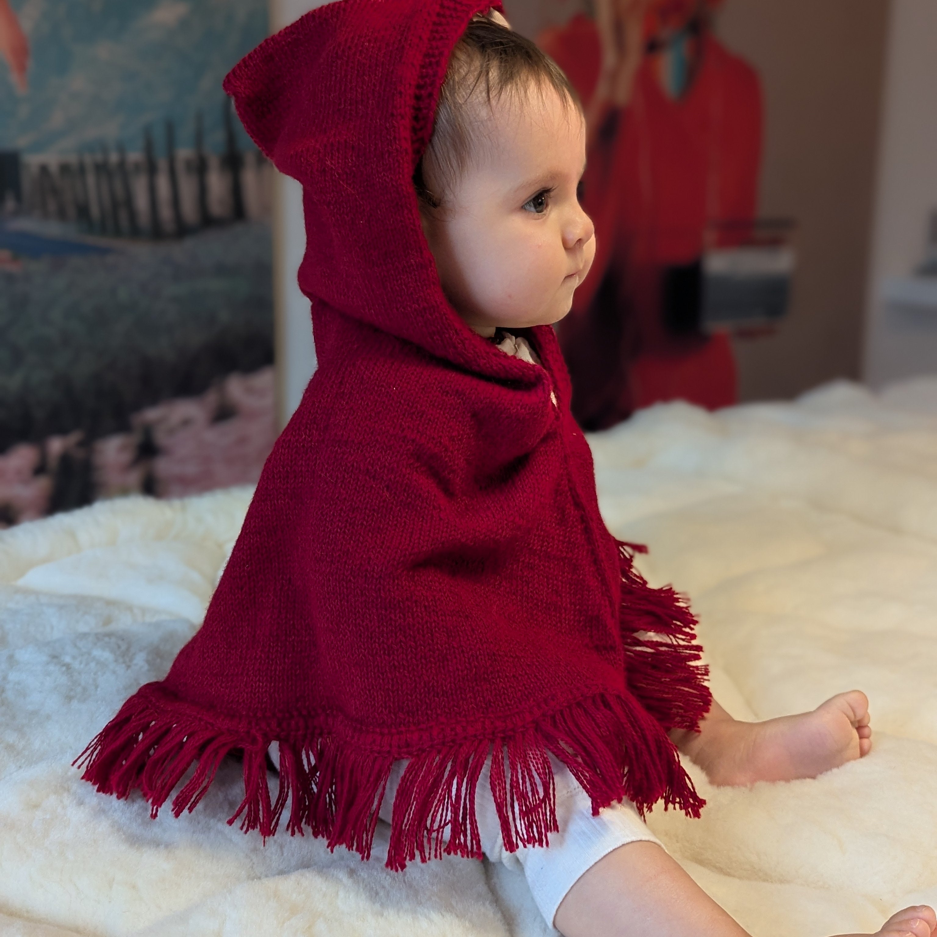Baby wearing a red hooded cloak sitting on a white surface with a colorful background.
