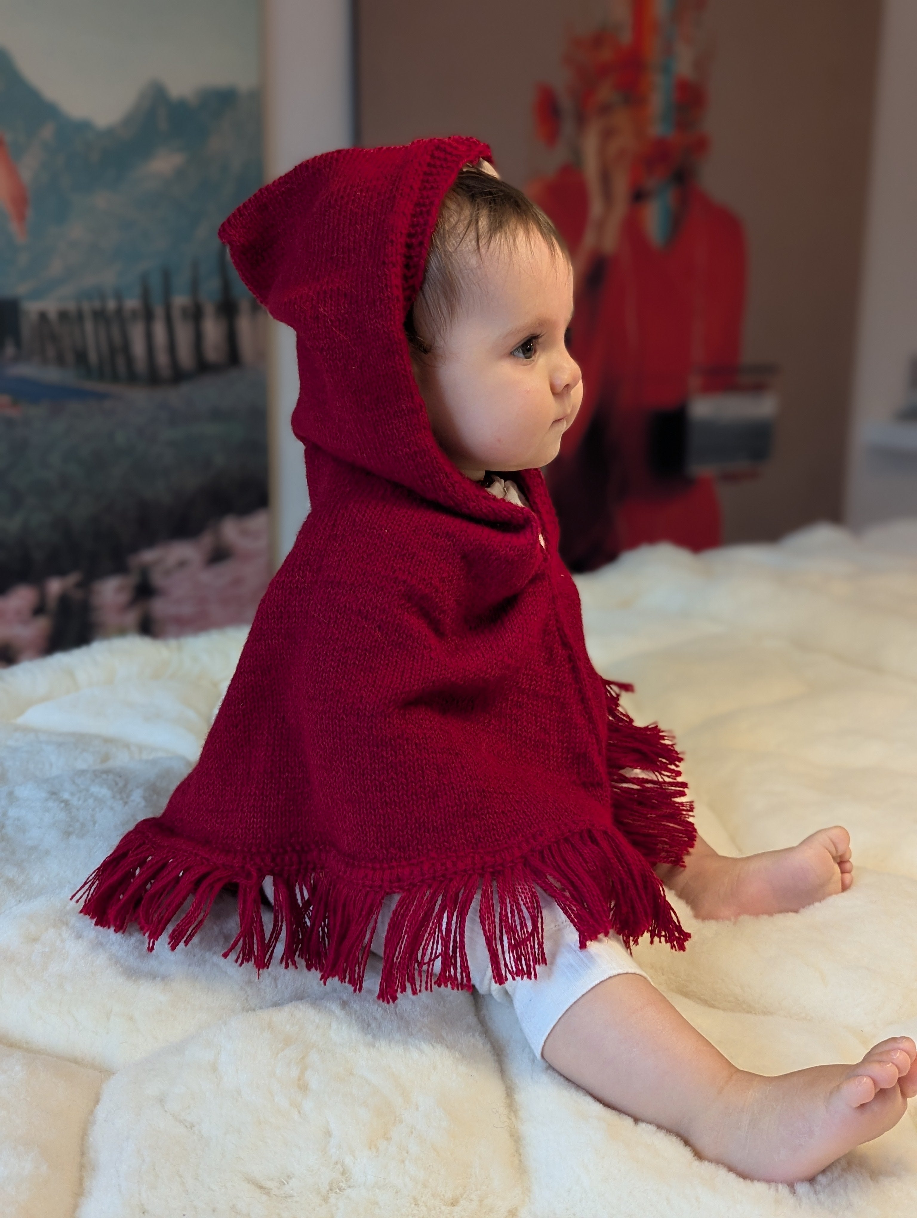 Baby wearing a red hooded cloak sitting on a white surface with a colorful background.