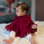 Baby wearing a red poncho sitting on a white blanket with a colorful mural in the background