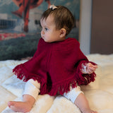Baby wearing a red poncho sitting on a white blanket with a colorful mural in the background