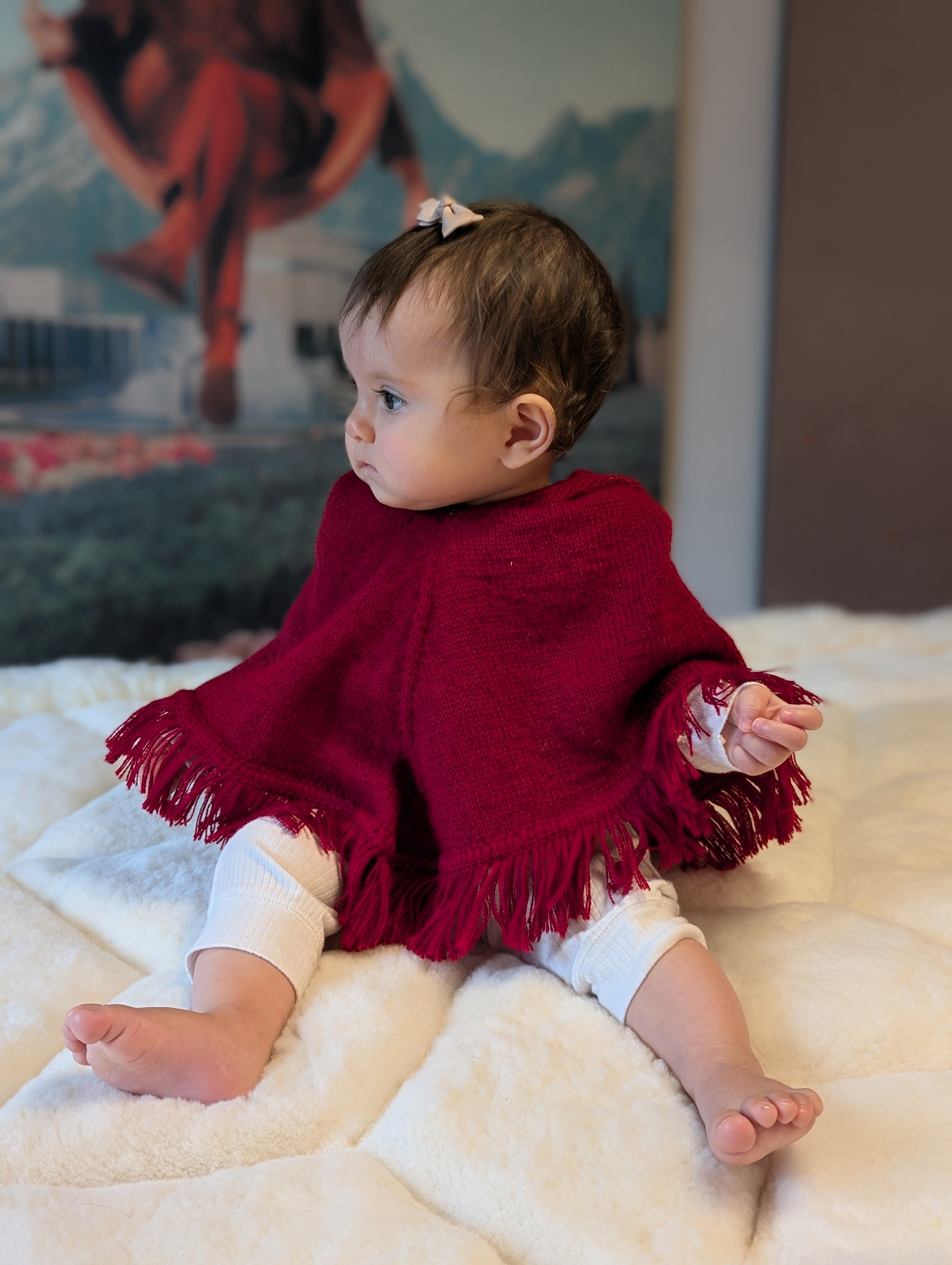 Baby wearing a red poncho sitting on a white blanket with a colorful mural in the background