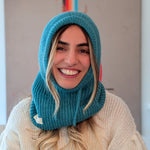 Woman wearing a teal knitted hood over her head, smiling indoors.