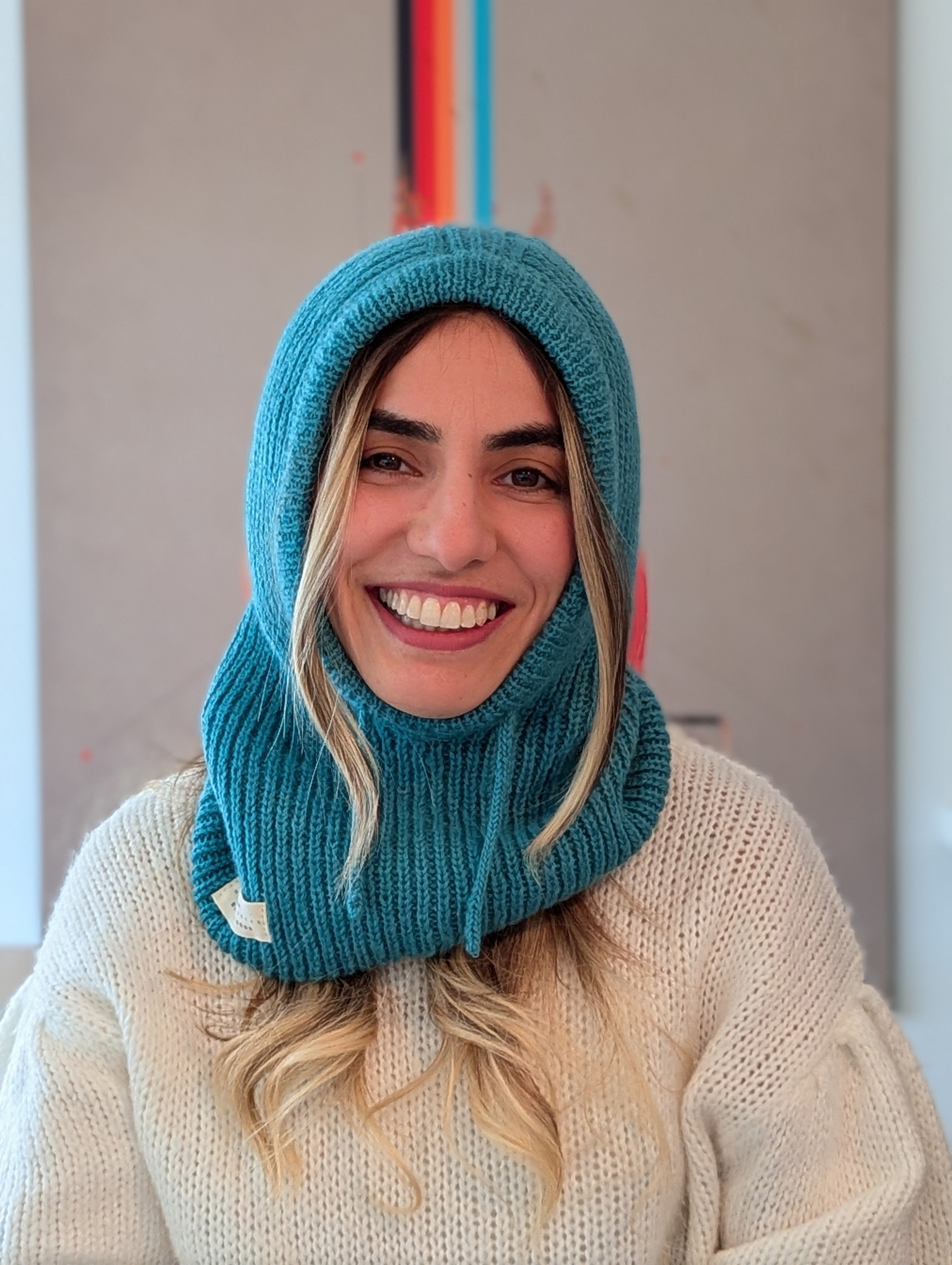 Woman wearing a teal knitted hood over her head, smiling indoors.